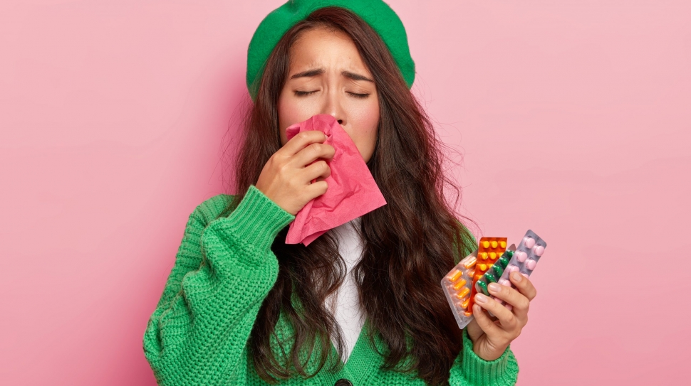 Unhappy brunette girl suffers from flu symptoms, rubs nose with handkerchief, has cold, holds pills, wears green jumper and cap, isolated on pink wall. Seasoanl disease, running nose and medicaments