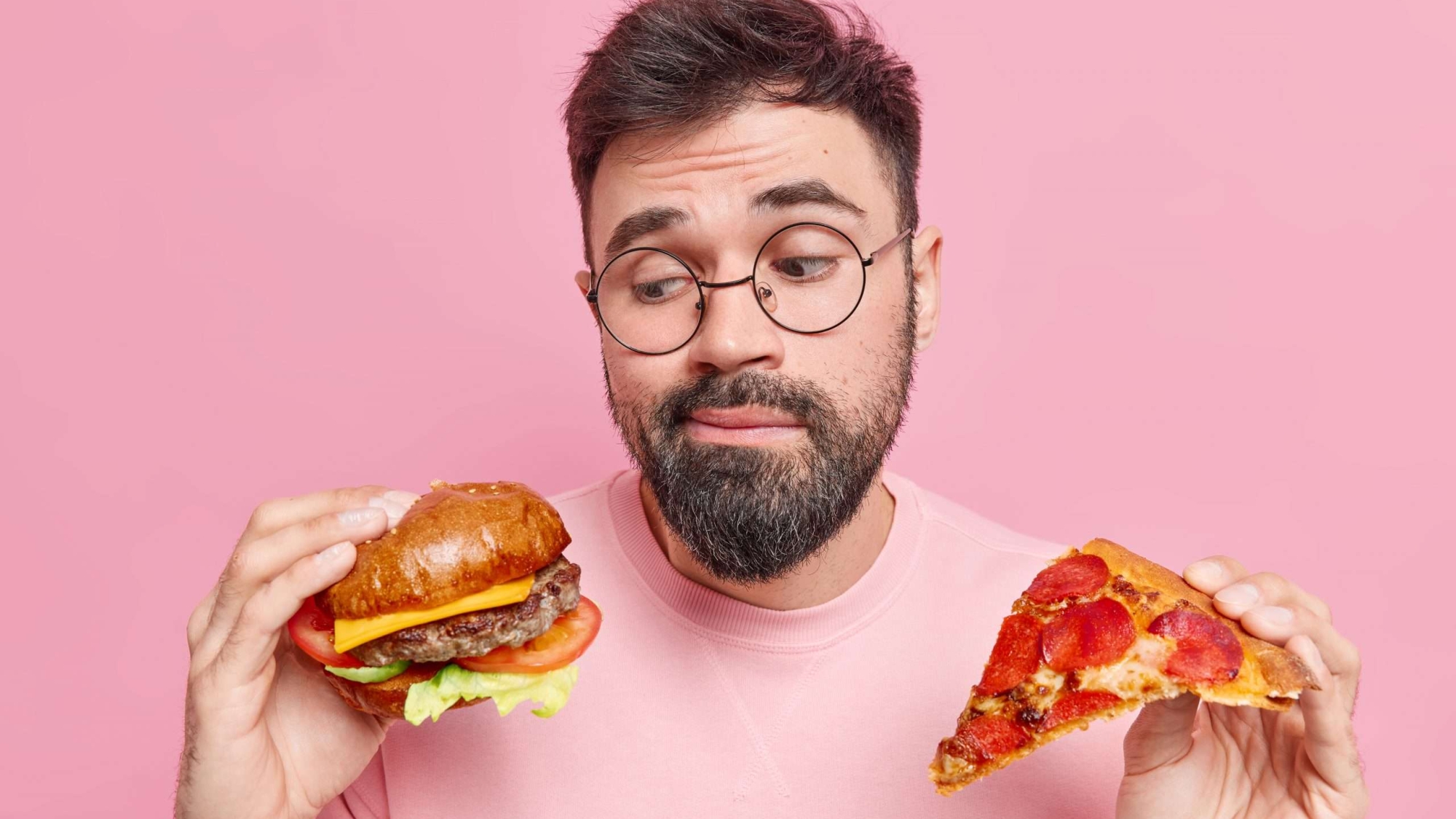 Handsome bearded adult European man feels hesitant whether to eat hamburger or pizza prefers eating junk food wears round spectacles and jumper isolated over pink background. Cheat meal concept