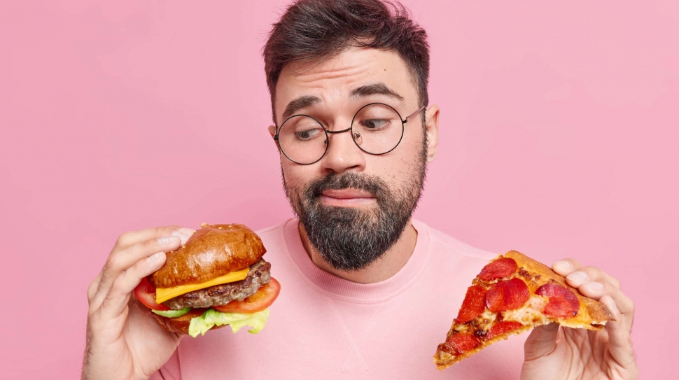 Handsome bearded adult European man feels hesitant whether to eat hamburger or pizza prefers eating junk food wears round spectacles and jumper isolated over pink background. Cheat meal concept