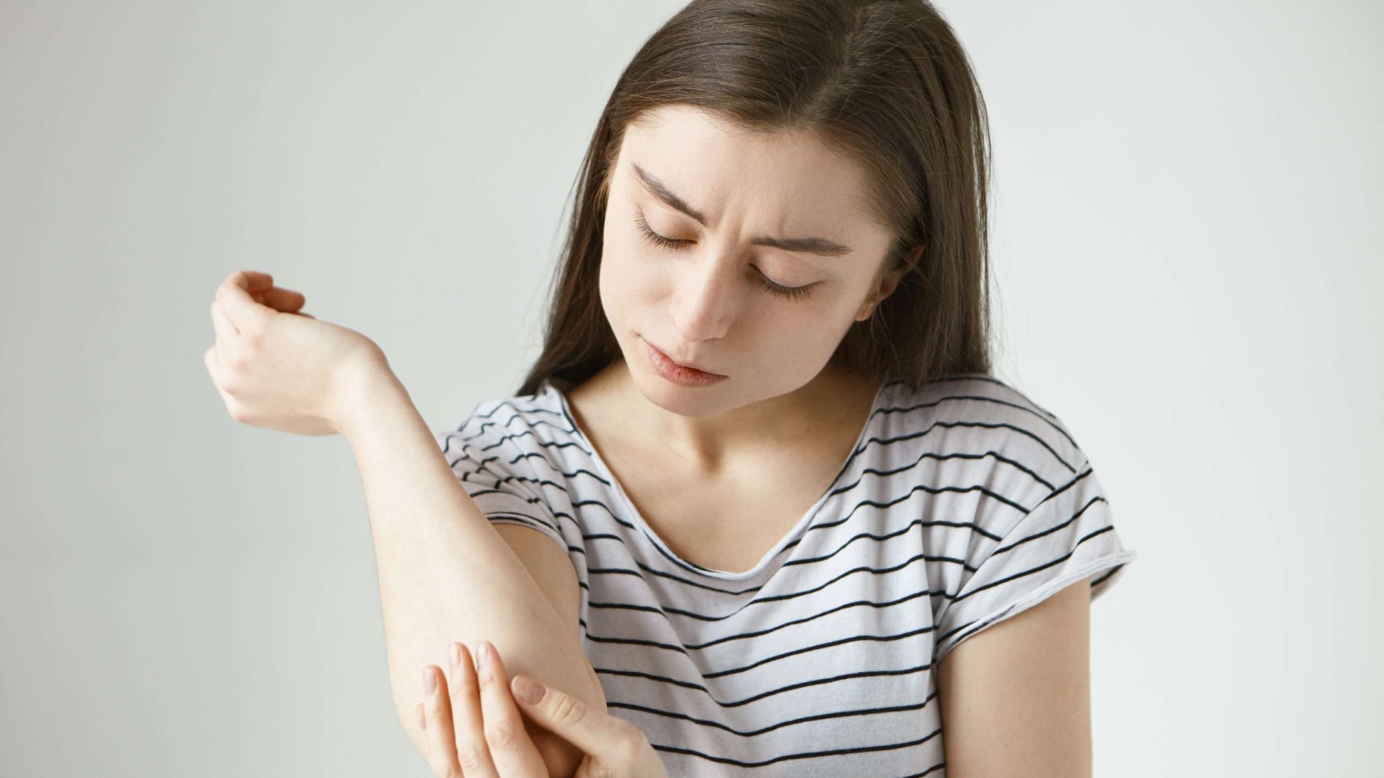 Studio picture of frustrated young dark haired female studying skin on her arm after she fell off bike. Student girl dressed in stylish striped t-shirt looking at her elbow, feeling itch or pain