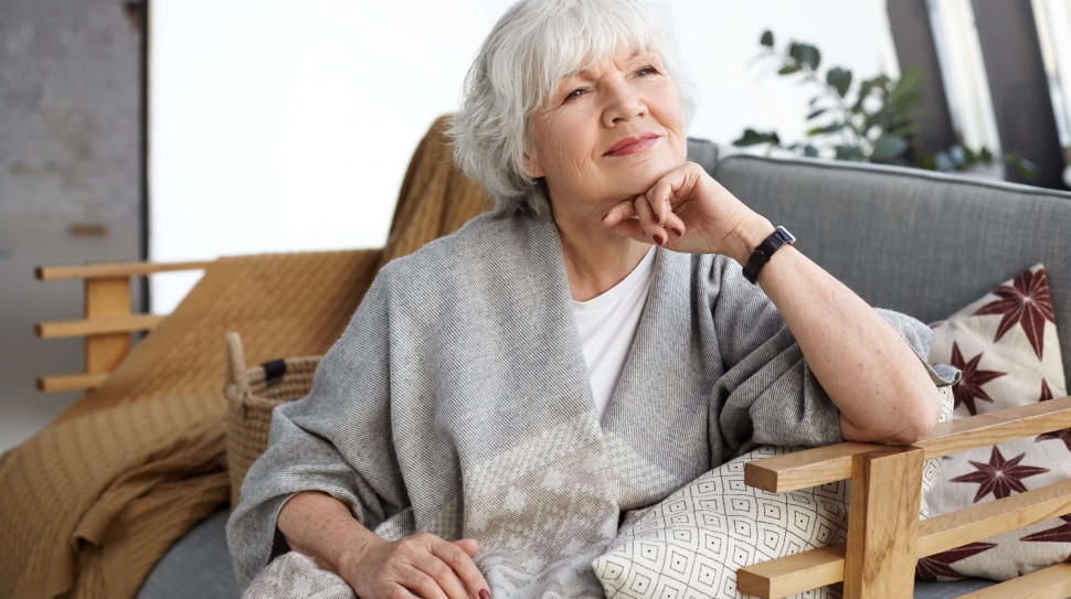Portrait of lovely middle aged grey haired European woman with dreamy smile and eyes full of wisdom relaxing at home alone, sitting on comfortable couch, reminiscing about days of her youth