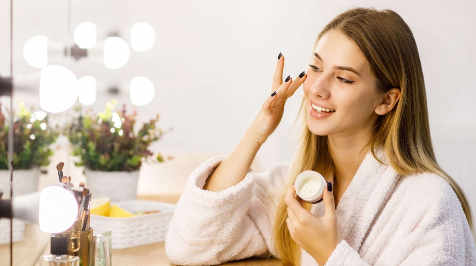 Young beautiful blonde girl cares for face skin with moisturizer in front of mirror.