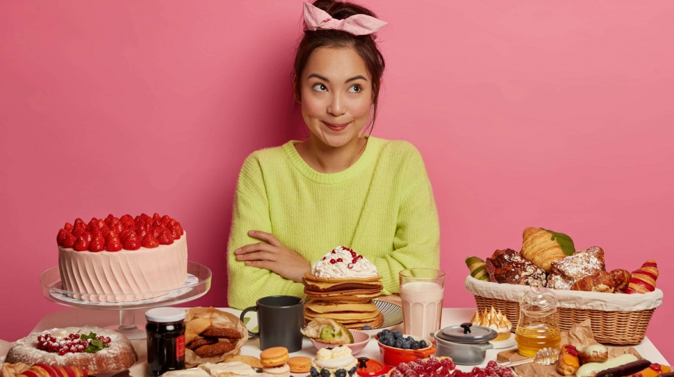 Pensive female sweettooth enjoys eating sweet desserts, poses at table full of tasty cakes, pancakes, cookies, drinks coffee or milk, surrounded by junk food containing much sugar. Culinary, baking